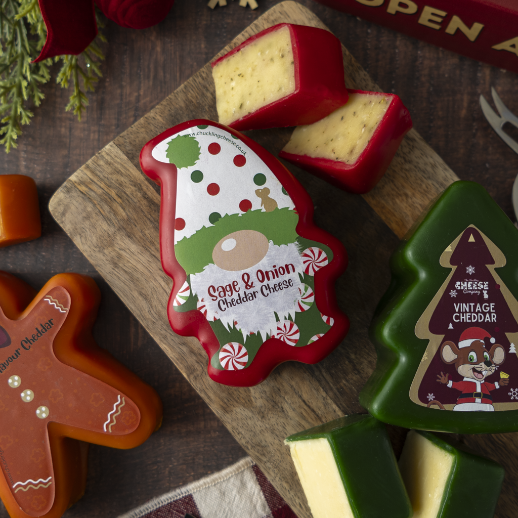 A styled holiday cheese assortment on a wooden board, featuring a Christmas gnome-shaped cheddar cheese labeled 'Sage & Onion Cheddar Cheese,' a green tree-shaped cheese labeled 'Vintage Cheddar,' and other seasonal cheese shapes around it.