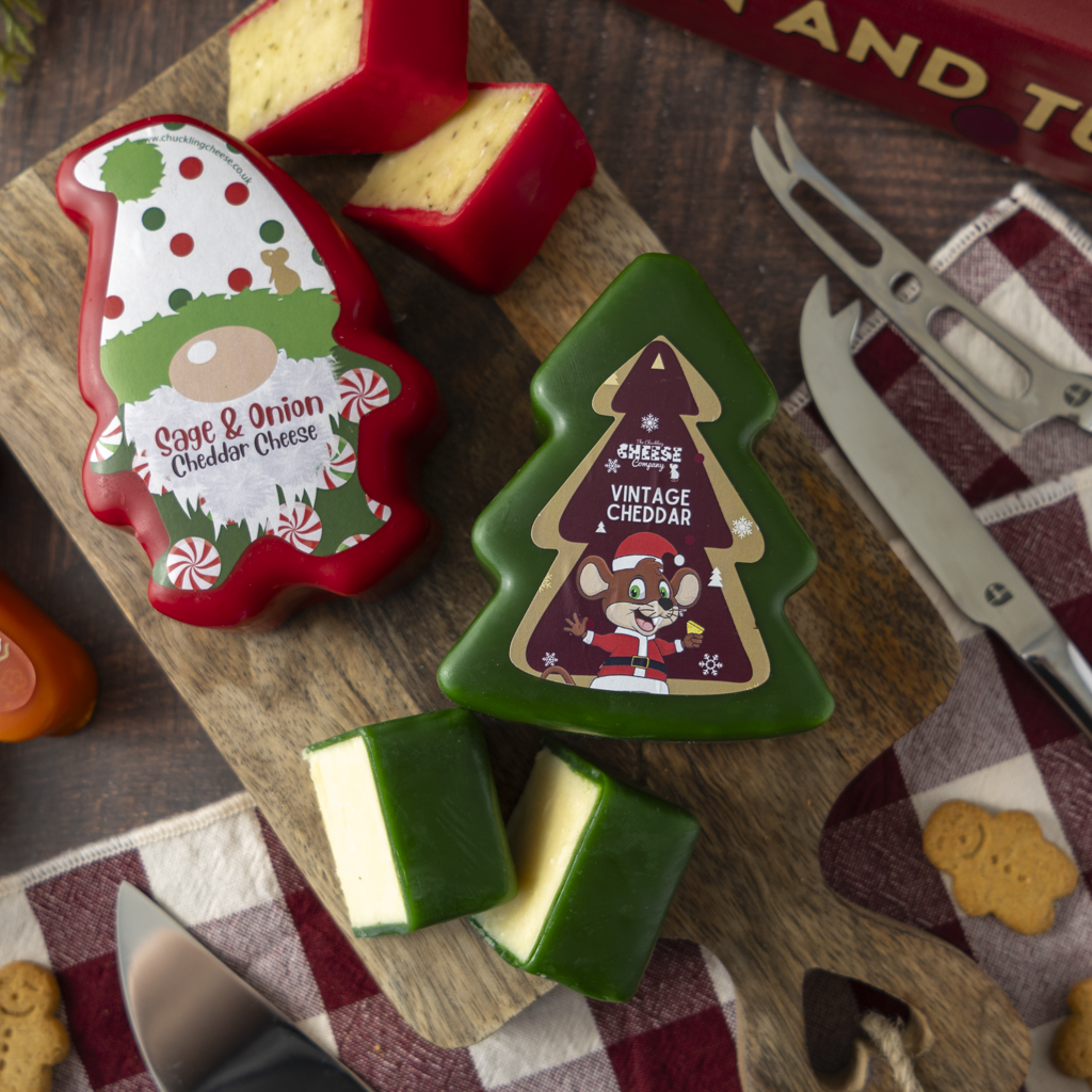 Two shaped cheese truckles on a wooden board: a red Christmas gnome cheese with white cheddar inside and a green Christmas tree cheese with white cheddar inside. Knives, a checkered cloth, and cookie props surround them on a dark wood surface.