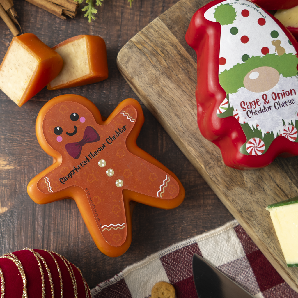 A styled flat-lay of cheeses on a dark wooden table, featuring a gingerbread man-shaped cheddar cheese slice in the foreground and a red Christmas-themed cheddar cheese package on a wooden board in the background.