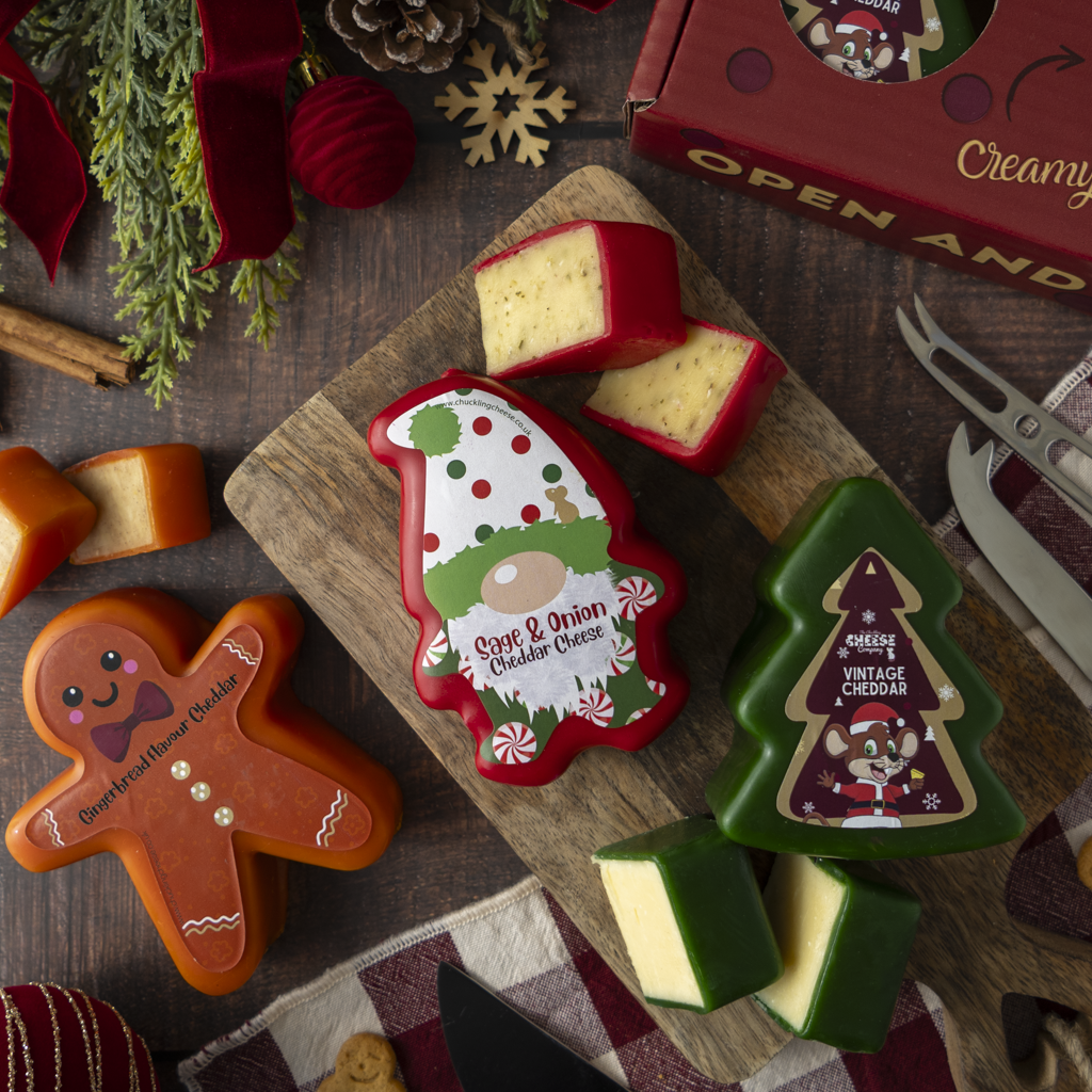 Christmas-themed cheese gift set featuring gingerbread man and tree-shaped cheddar cheeses on a wooden cutting board, with a red gift box and holiday decorations in the background.