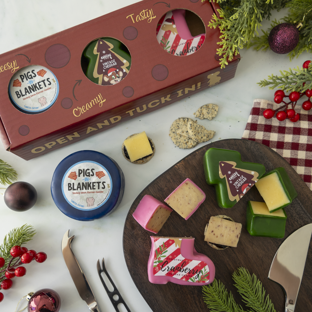 A styled holiday cheese and crackers spread featuring a red gift box labeled 'Pigs in Blankets' with round and shaped cheese pieces, a round blue-labeled cheese, crackers, knives, pine branches, red berries, and a red check cloth on a white surface.