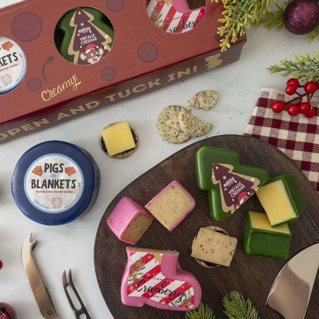 A festive cheese and snack assortment styled on a white surface with a red gift box, a round blue-labled cheese called Pigs in Blankets, and several cheese wedges and crackers arranged on a dark wooden board. The scene includes holiday greenery and a red-and-white check cloth.