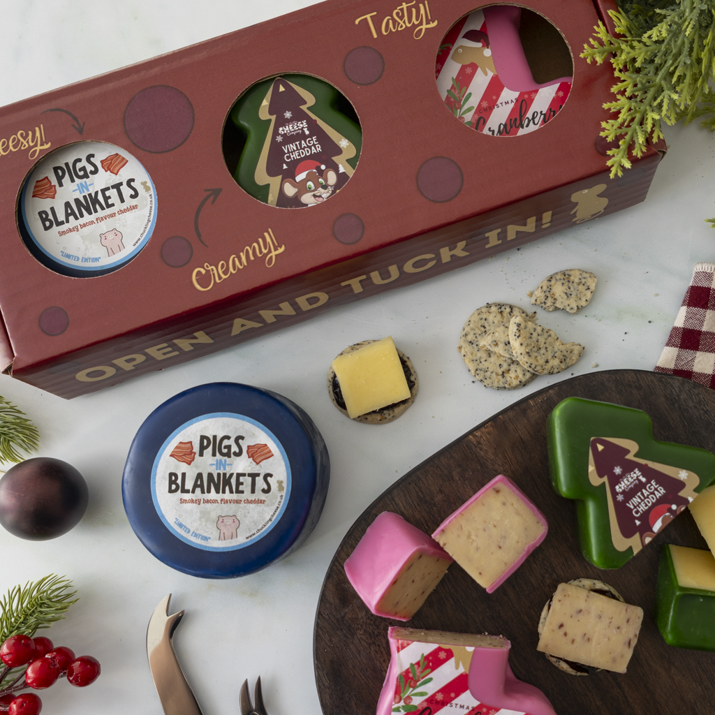 A holiday gift set from The Chuckling Cheese Company arranged on a white surface, featuring a red gift box with circular cutouts, a round blue-wrap cheese truckle, cheese wedges and slices on a wooden board, cookies, and festive greenery. Visible branding reads Pigs in  Blankets.