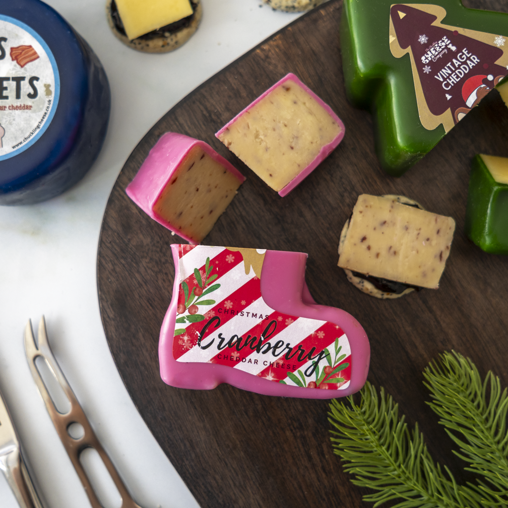 A styled cheese board scene with a pink boot-shaped cheese labeled Cranberry Cheese, alongside other cheeses and serving tools on a dark wooden board.