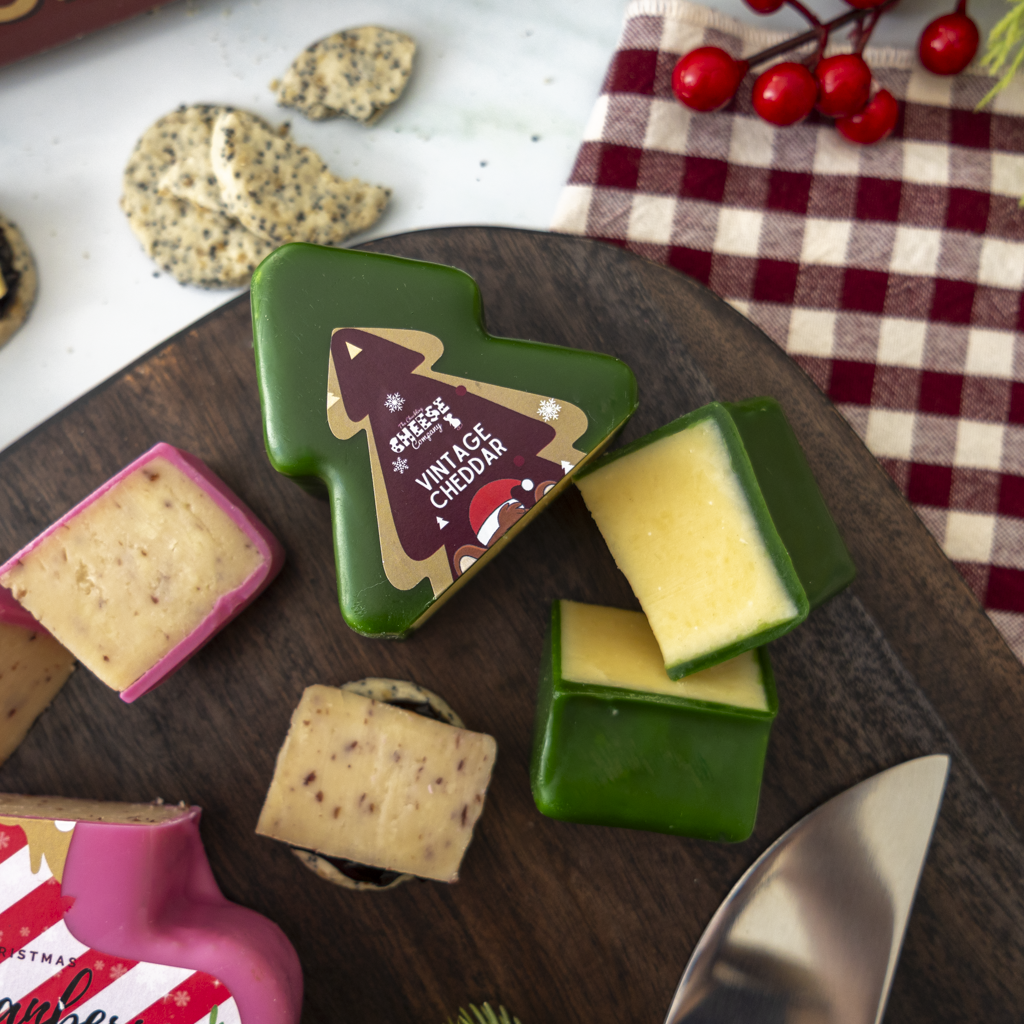 A styled food product shot showing several Christmas-themed cheese wheels and wedges on a dark wooden board. The cheeses have green, pink, and pale yellow rinds with cream-coloured cheddar centers.