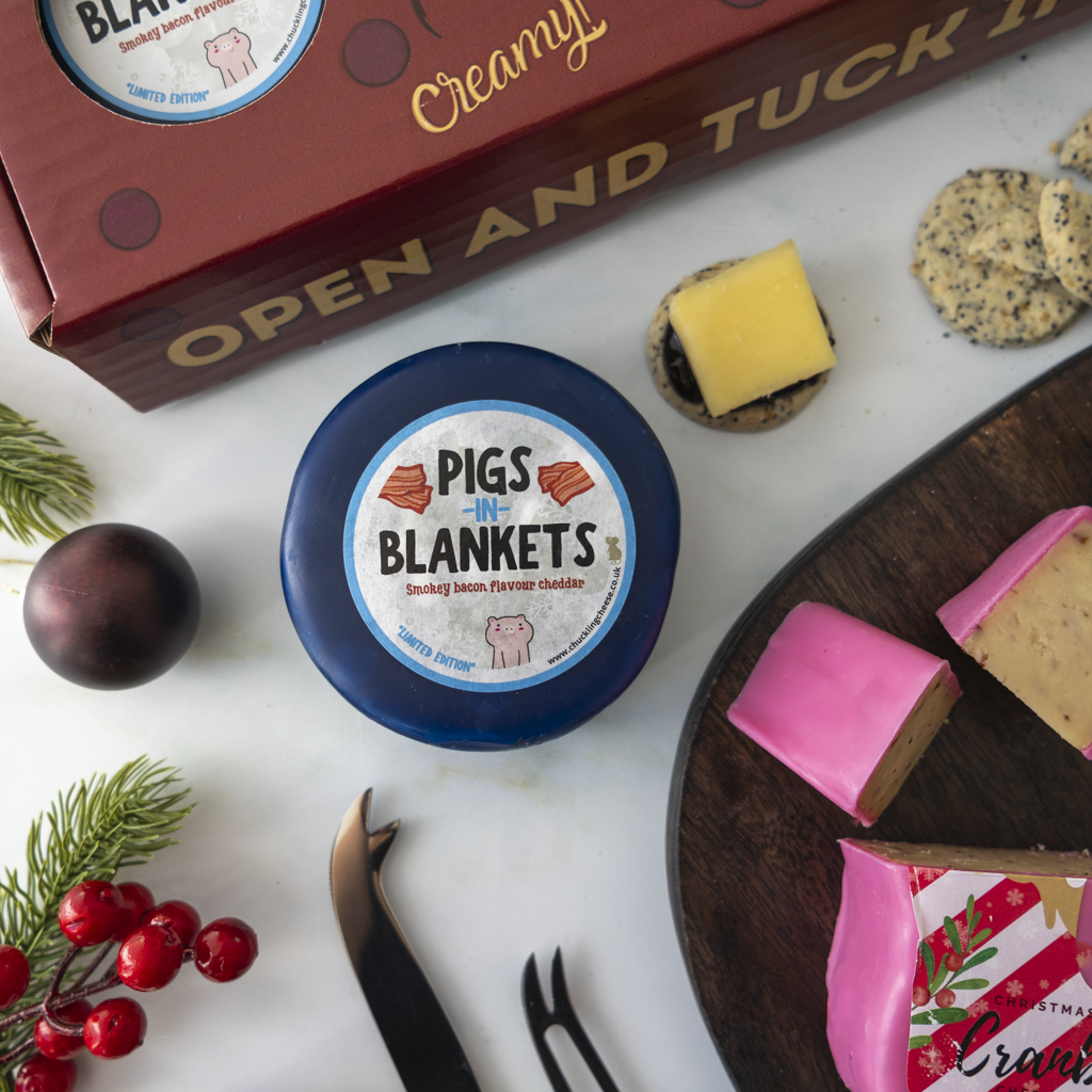 A styled flat-lay featuring a round blue cheese labeled 'Pigs in Blankets' in the center, with a red gift box, cheese wedges, crackers, and holiday decorations arranged around it on a white surface.