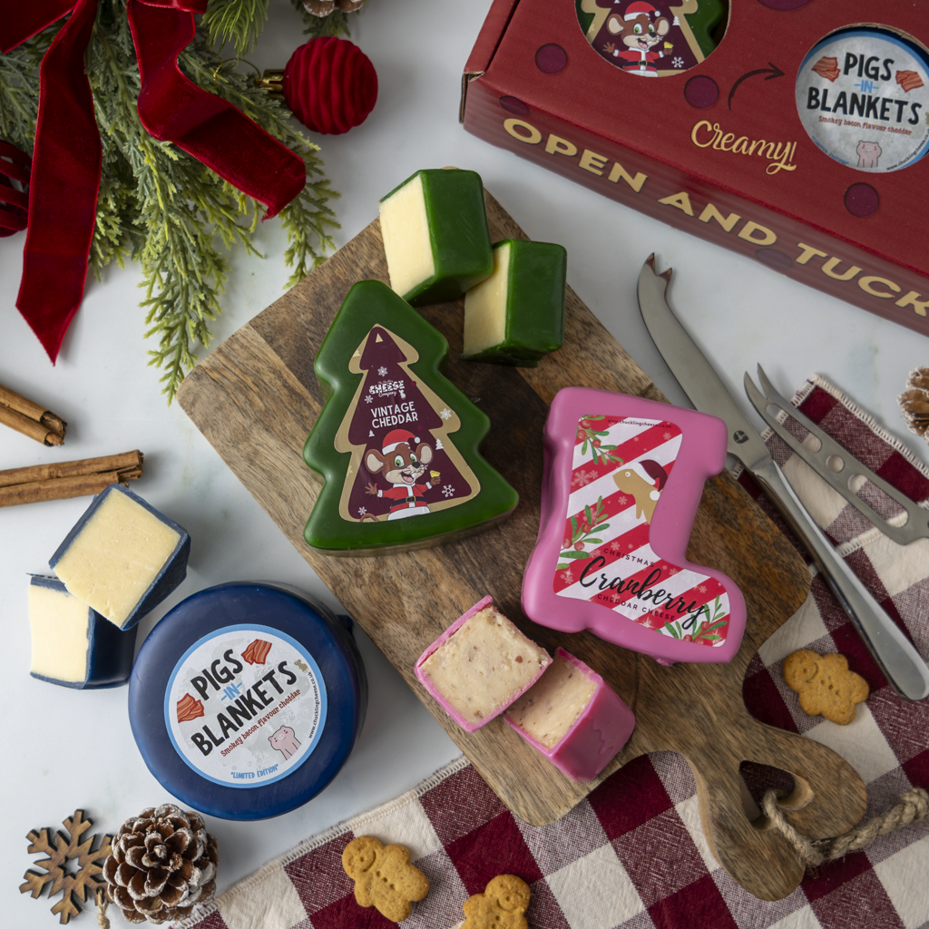 A styled flat-lay of Christmas cheese products on a wooden board with holiday decorations. Visible items include a Christmas tree-shaped cheese labelled 'Vintage Cheddar', a pink boot-shaped cheese labelled 'Cranberry', a round blue cheese container labelled 'Pigs in Blankets', wedges of pale cheese, a red gift box, and serving tools on a white background.