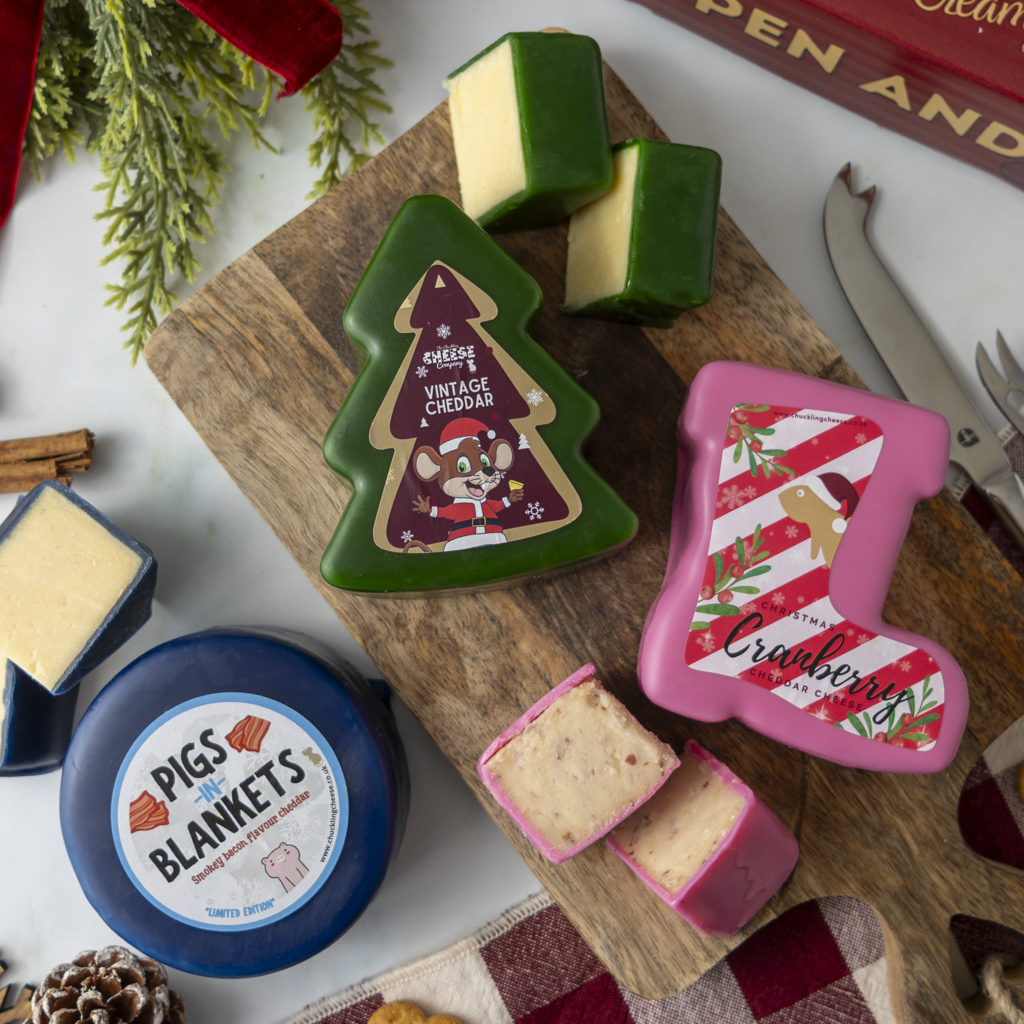 A holiday-themed cheese board display featuring a green Christmas tree-shaped cheddar cheese with a 'Merry Vintage Cheddar' label, a pink stocking-shaped cranberry cheddar with a 'Cranberry' label, a round blue 'Pigs in Blankets' cheese, and other cheeses on a wooden cutting board with festive props.
