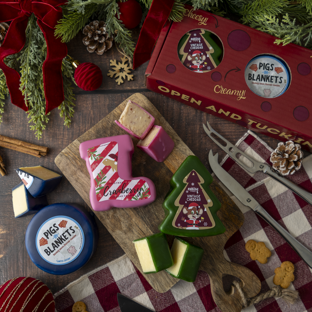 A styled flat lay of Christmas-themed cheese truckles and packaging on a wooden board with carving tools, surrounded by holiday greenery and red ribbon.