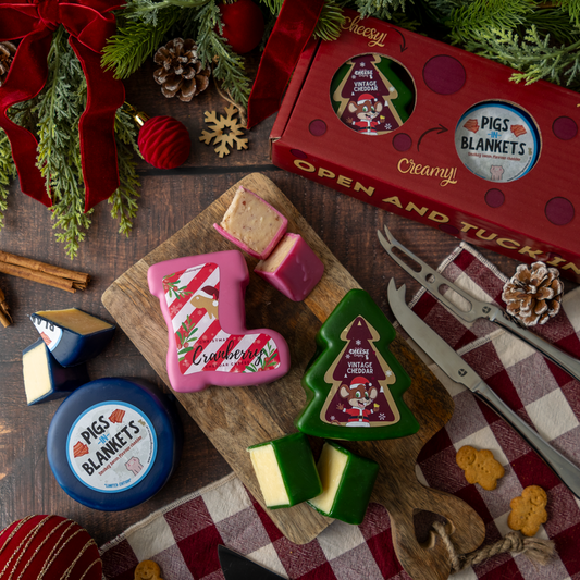 A styled flat lay of Christmas-themed cheese truckles and packaging on a wooden board with carving tools, surrounded by holiday greenery and red ribbon.