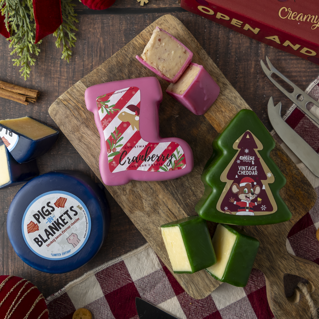 A styled holiday cheese assortment on a wooden board, including a pink Christmas stocking-shaped cheese labeled Cranberry and a green Christmas tree-shaped cheese labeled Vintage Cheddar, with cut wedges showing the pale cheese interior. A round blue container labeled Pigs Blankets is also visible, along with cheese knives and a red checkered cloth on a dark wood surface.
