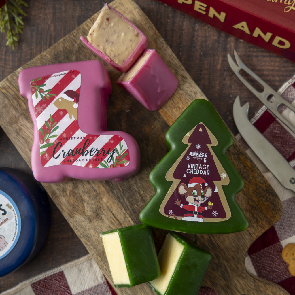 Holiday-themed cheese blocks shaped like a Christmas stocking and a tree, displayed on a wooden board with wedges cut from the cheese. The stocking cheese is pink with a red-and-white striped holiday label reading Cranberry Cheddar, and the tree cheese is green with a vintage cheddar label.