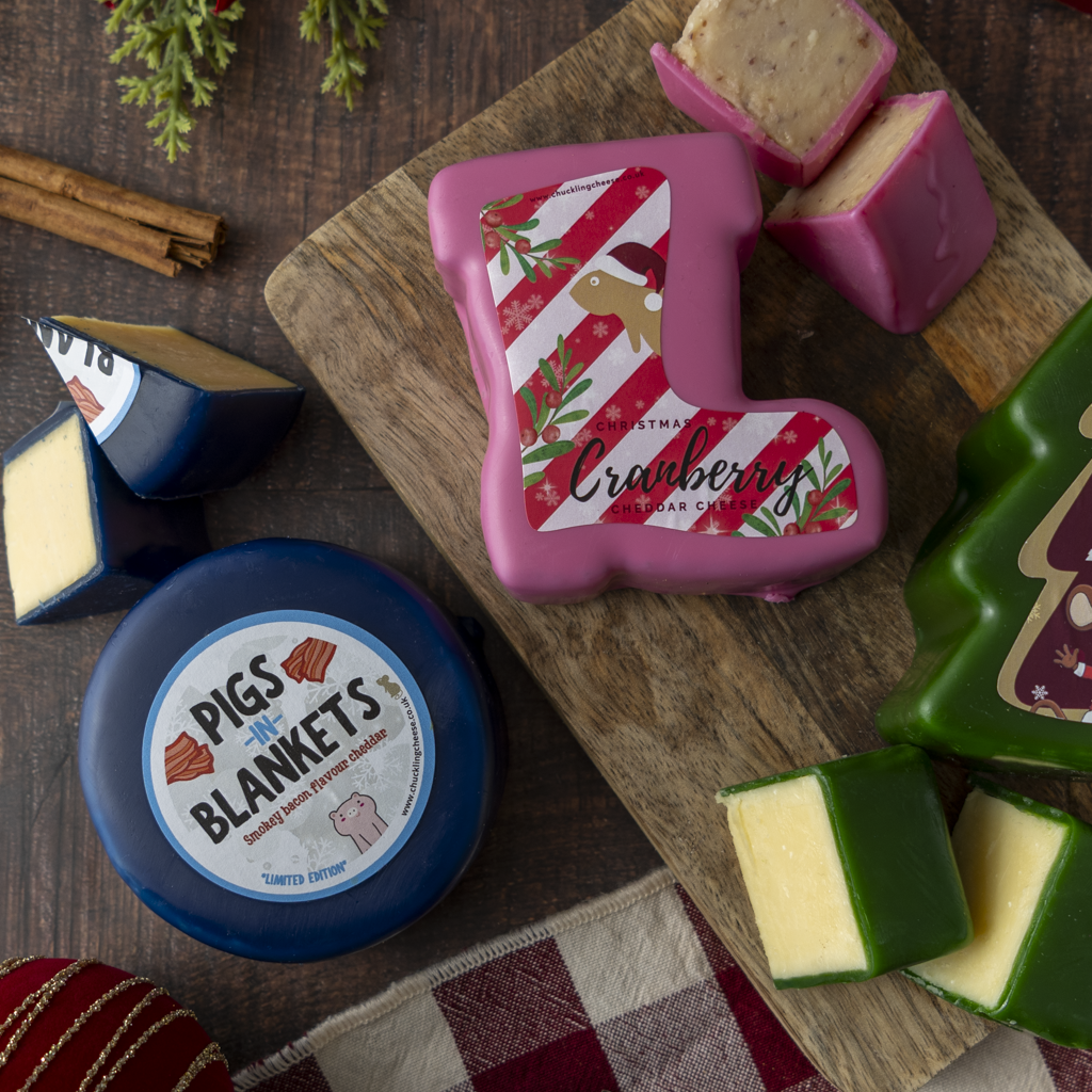 A styled flat lay of gourmet cheese wheels and wedges on a dark, rustic wooden table. The main featured product is a Christmas stocking-shaped cheese wheel with a pink, red, and white striped holiday label reading 'Cranberry Cheddar Cheese'.