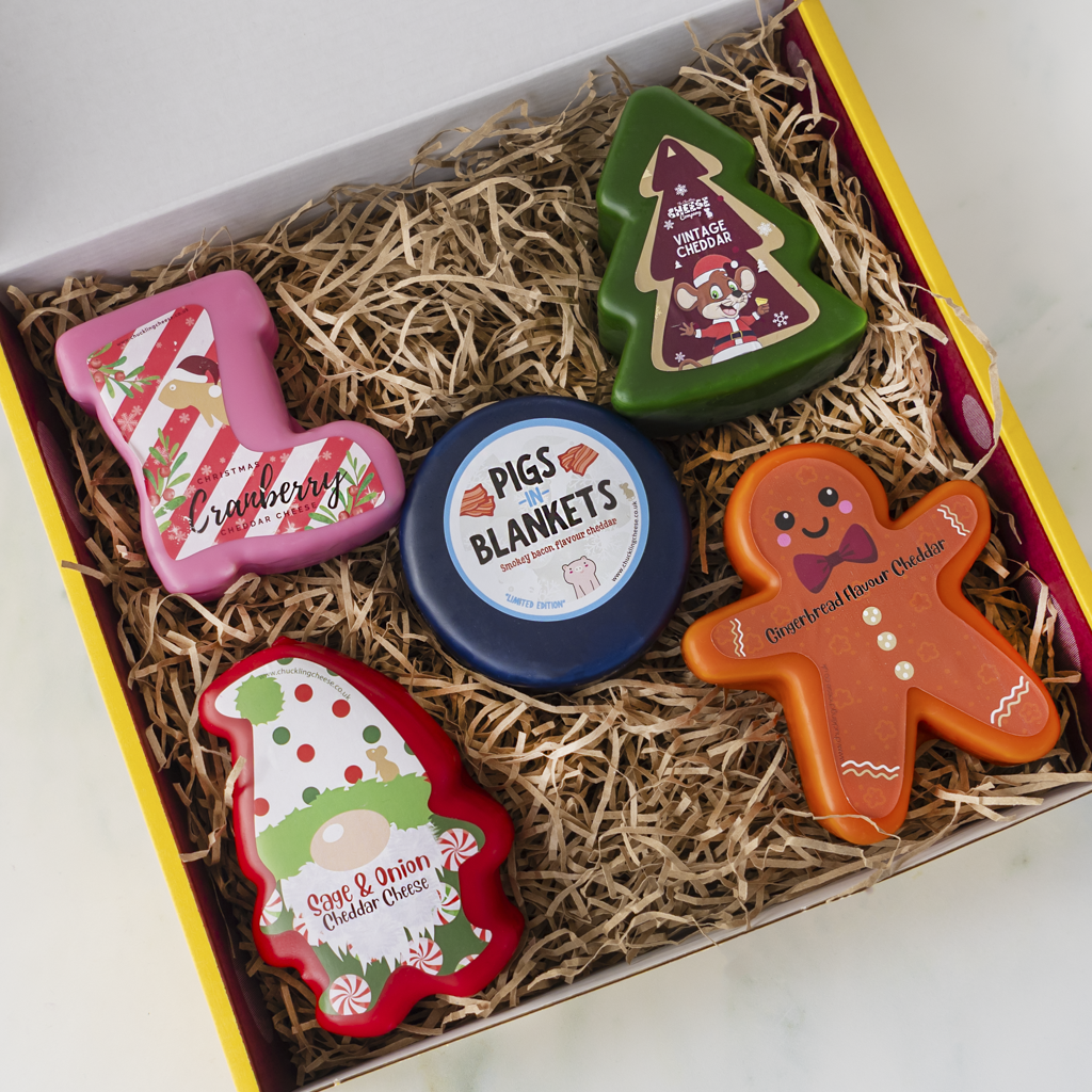 A Christmas-themed gift box filled with shredded brown paper, containing five small decorative cheese truckles: a pink stocking-shaped item labelled 'Cranberry', a green Christmas tree-shaped item labelled 'Vintage Cheddar', a round blue tin labelled 'Pigs in Blankets', an orange gingerbread man-shaped item labelled 'Gingerbread flavour cheddar', and a red gnome-shaped item labelled 'Sage & Onion Cheddar Cheese'.