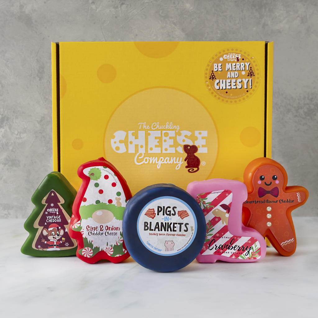 A gift set from The Chuckling Cheese Company featuring a yellow branded box and five novelty cheese truckles shaped like Christmas trees, Sage and Onion, a pigs in blankets, a cranberry, and a gingerbread man, displayed on a white marble surface against a grey textured background.