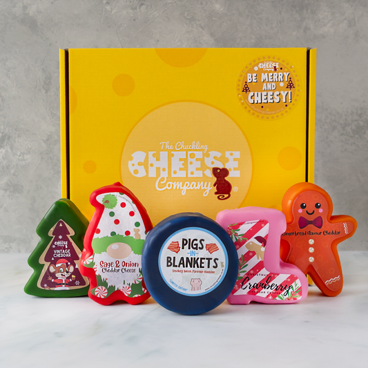 A gift set from The Chuckling Cheese Company featuring a yellow branded box and five novelty cheese truckles shaped like Christmas trees, Sage and Onion, a pigs in blankets, a cranberry, and a gingerbread man, displayed on a white marble surface against a grey textured background.