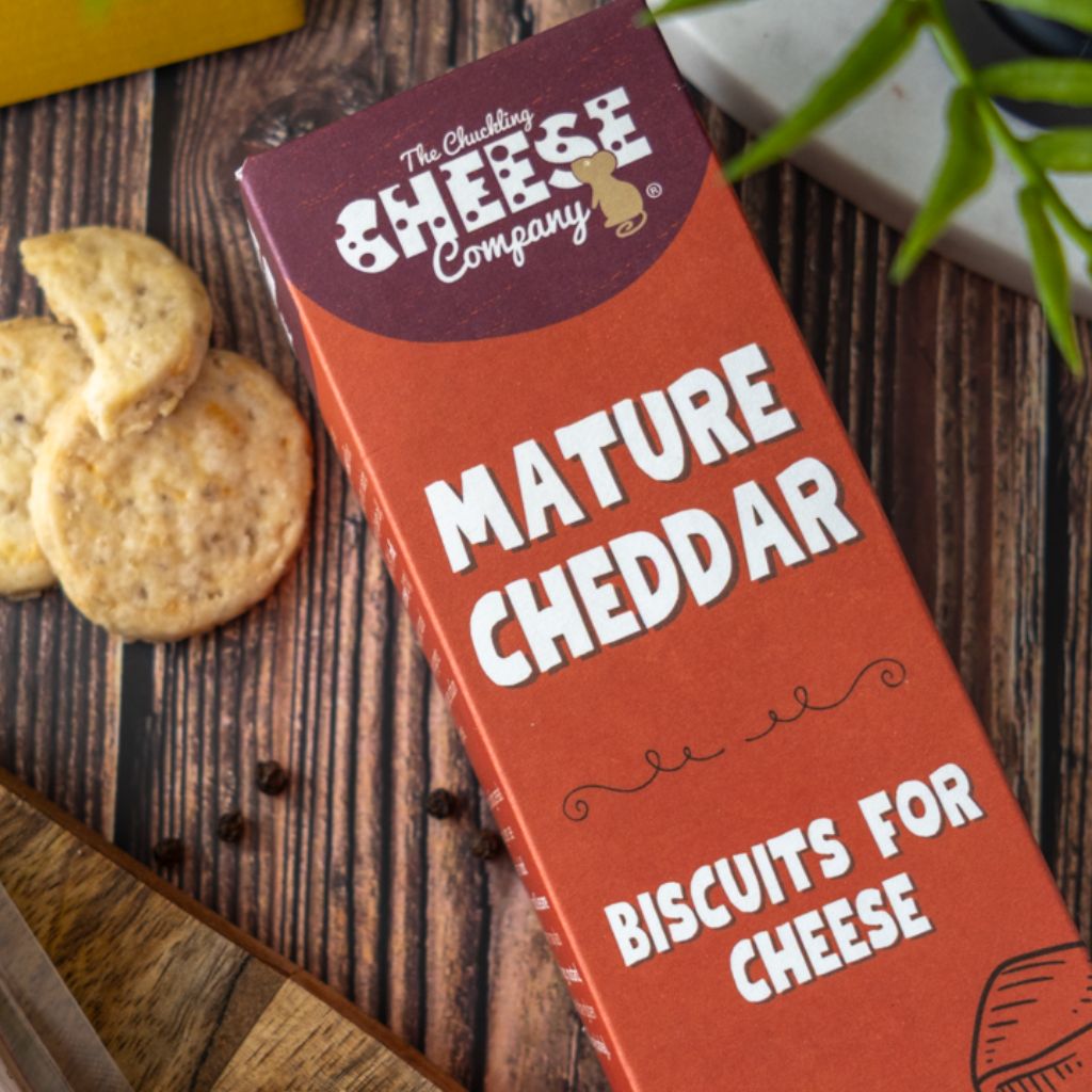 A package of The Chuckling Cheese Company Mature Cheddar Biscuits For Cheese, shown on a rustic wooden table next to some cheddar biscuits.