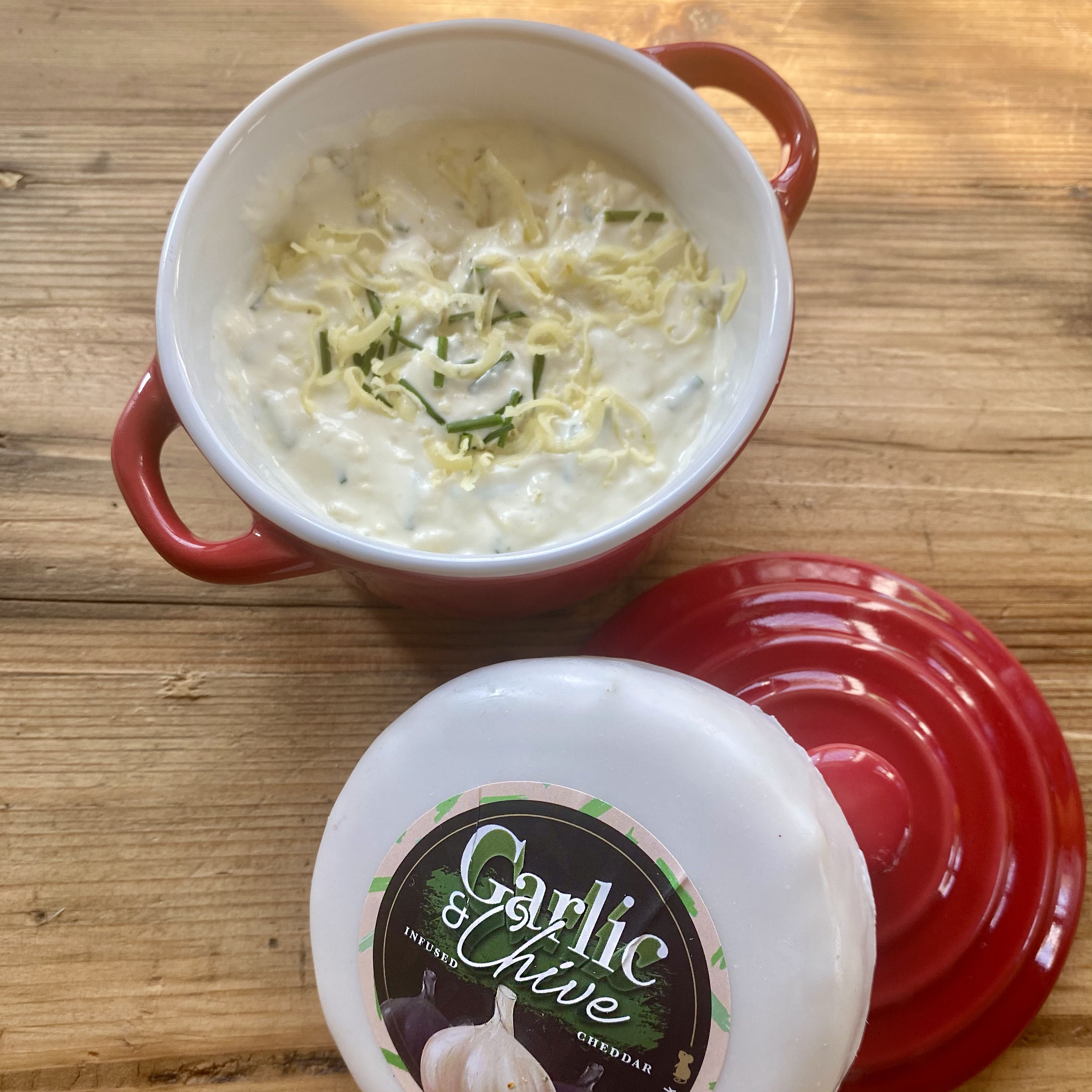 Red ramakin with creamy, chunky garlic and chive cheese dip with the white Garlic and Chive cheese truckle stood upon the ramakin lid.