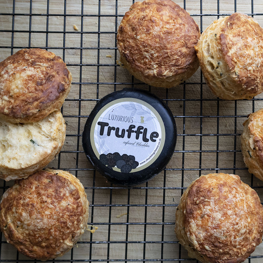 Golden cheese scones spread across a wire cooling tray. In the centre is the Truffle Cheese wax truckle packaging. One cheese scone on the left has been opened, showing the heavy flecks of me