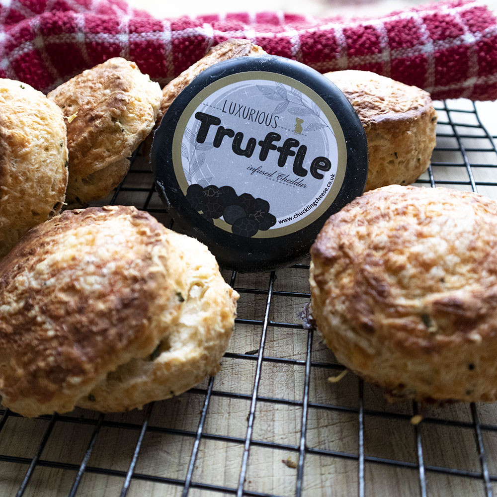 Golden cheese scones spread across a wire cooling tray with a red tea towel in the background. One cheese scone on the left has been opened, showing the heavy flecks of truffle and lashings o