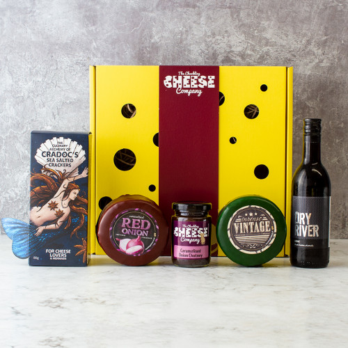 Grey background image of the W Cheese and Wine Gift Box including two cheese truckles, mini chutney, crackers, and small bottle of red wine.