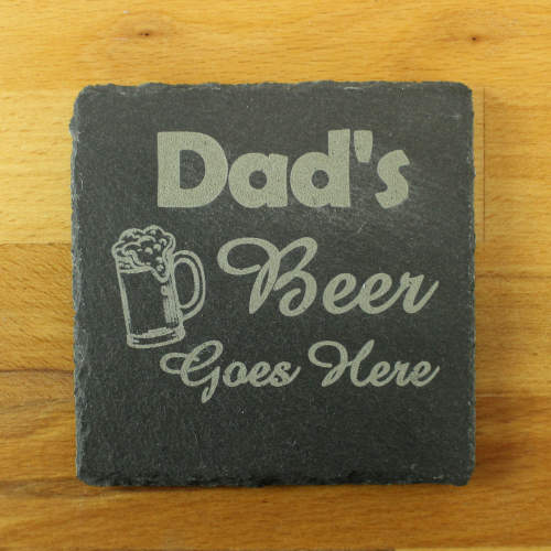 A square slate coaster by The Chuckling Cheese Company with 'Dad's Beer Goes Here' slogan engraved on it.