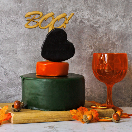 A Lifestyle Product Image Of The Halloween Celebration Cheese Cake Assembled And Rested On A Cheeseboard Decorated With Autumn Leafs And Nuts Topped With A Boo Cake Topper And Accompanied Wit