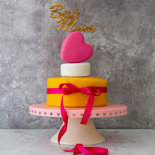 A Lifestyle Product Image Of Mums The Best Celebration Cheese Cake Assembled And Rested On A Cake Stand Tied With A Pink Ribbon And Topped With Best Mum Wooden Cake topper Presented On A Grey