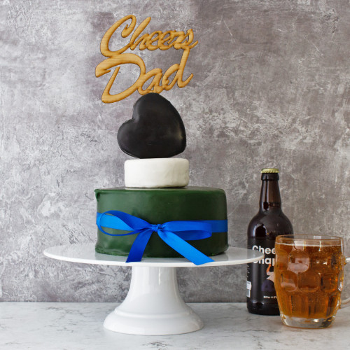 A lifestyle product image of Dad's The Best Celebration Cheese Cake assembled and rested on a cake stand, tied with a blue ribbon, topped with Cheers Dad cake topper. Available at The Chuckli