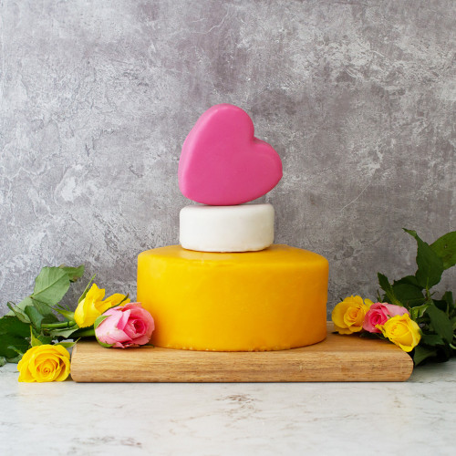 A Lifestyle Product Image Of The Chuckles Celebration Cheese Cake Assembled And Rested On A Cheeseboard Decorated With Yellow And Pink Roses Presented On A Grey Background. 