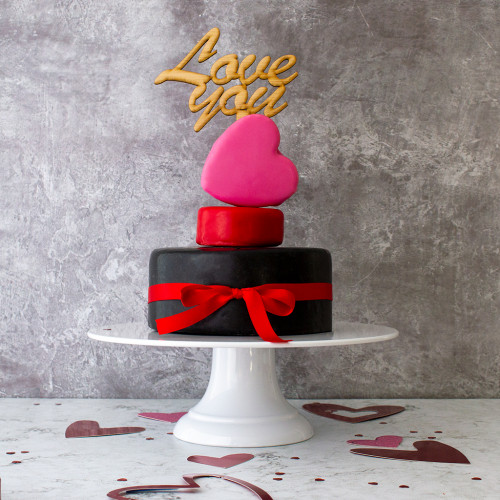 A Lifestyle Product Image Of The I Love You Celebration Cheese Cake Assembled And Rested On A Cake Stand Decorated With Love Hearts, Tied With A Red Ribbon And Topped With Love You Cake Toppe