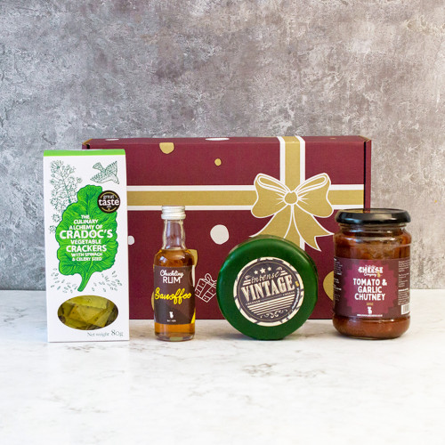 Grey background image of the Rum & Cheese Small Gift Box with tomato & garlic chutney, vintage cheddar truckle, 5cl Banoffee Rum, and a box of vegetable crackers.