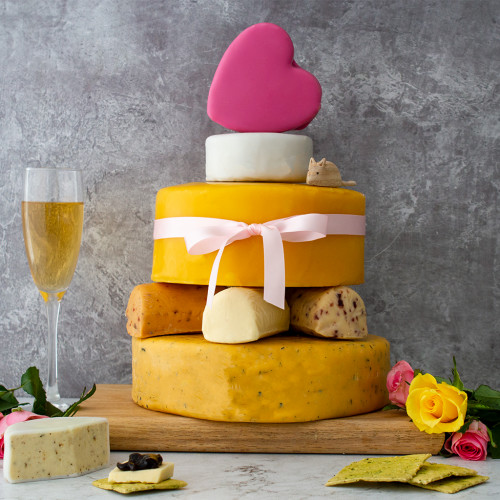 A Lifestyle Product Image Of The All Rounder Celebration Cheese Cake Assembled And Rested On A Cheeseboard Decorated With Yellow And Red Roses, Tied With A White Ribbon And Accompanied With A